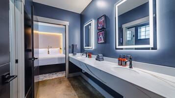 Une salle de bain moderne avec un long comptoir blanc, un grand miroir et un lit visible à travers une porte ouverte.