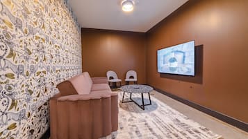 A room with a brown wall, a large screen, a brown sofa, two chairs, and a patterned rug.