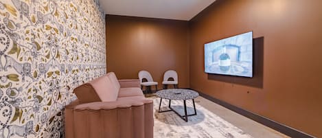 A room with a brown wall, a large screen, a brown sofa, two chairs, and a patterned rug.