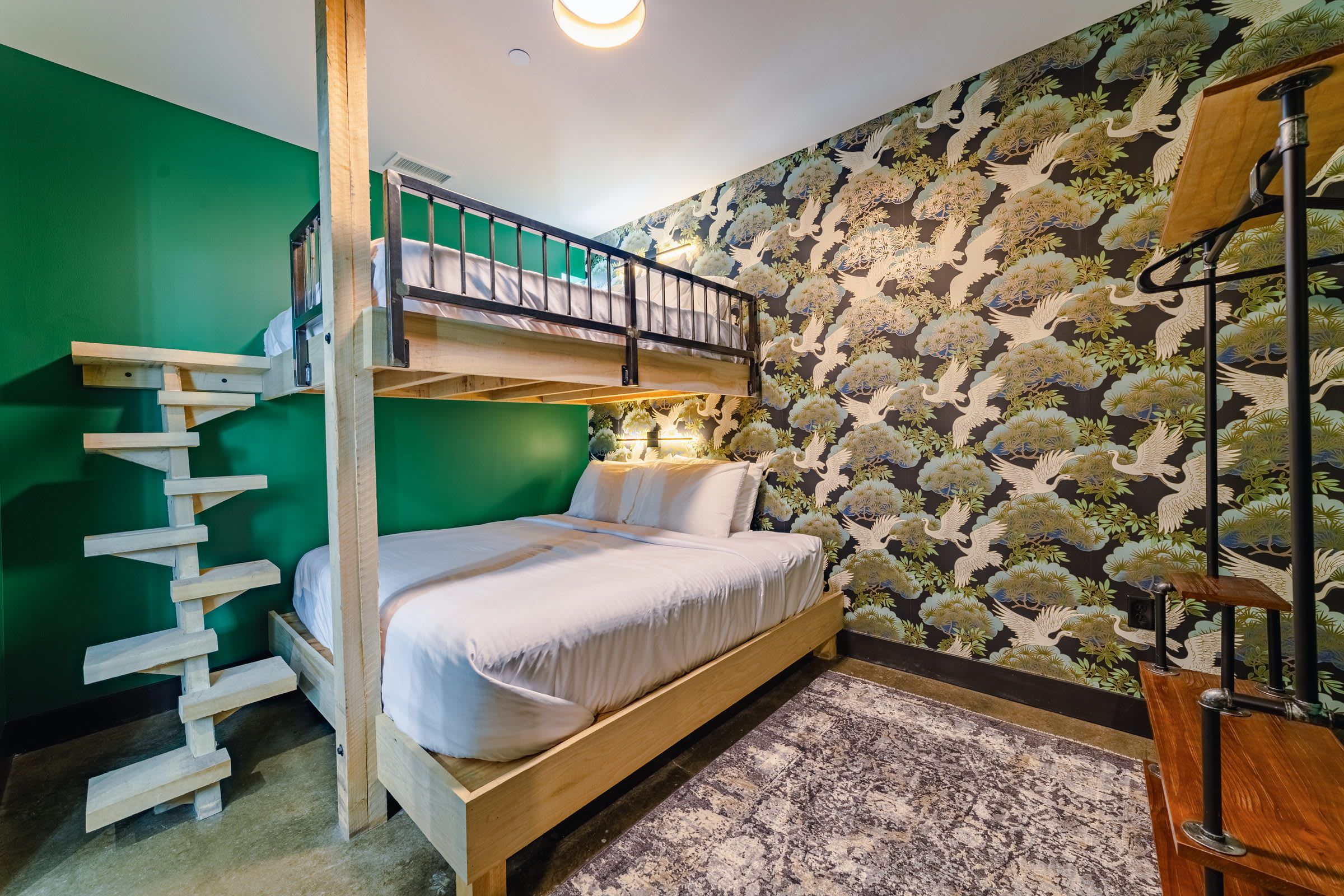 Quarto com beliche, escada de madeira, uma cama de solteiro com roupa de cama branca, parede estampada e uma mesinha com abajur.