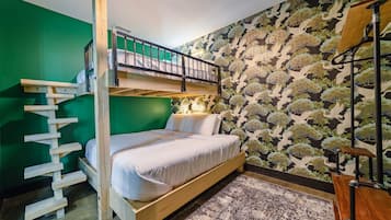 A bunk bed room with a wooden ladder, a single bed with white bedding, a patterned wall, and a small table with a lamp.