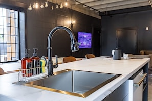 A modern kitchen with a large island, stainless steel sink, and a flat-screen TV mounted on the wall.