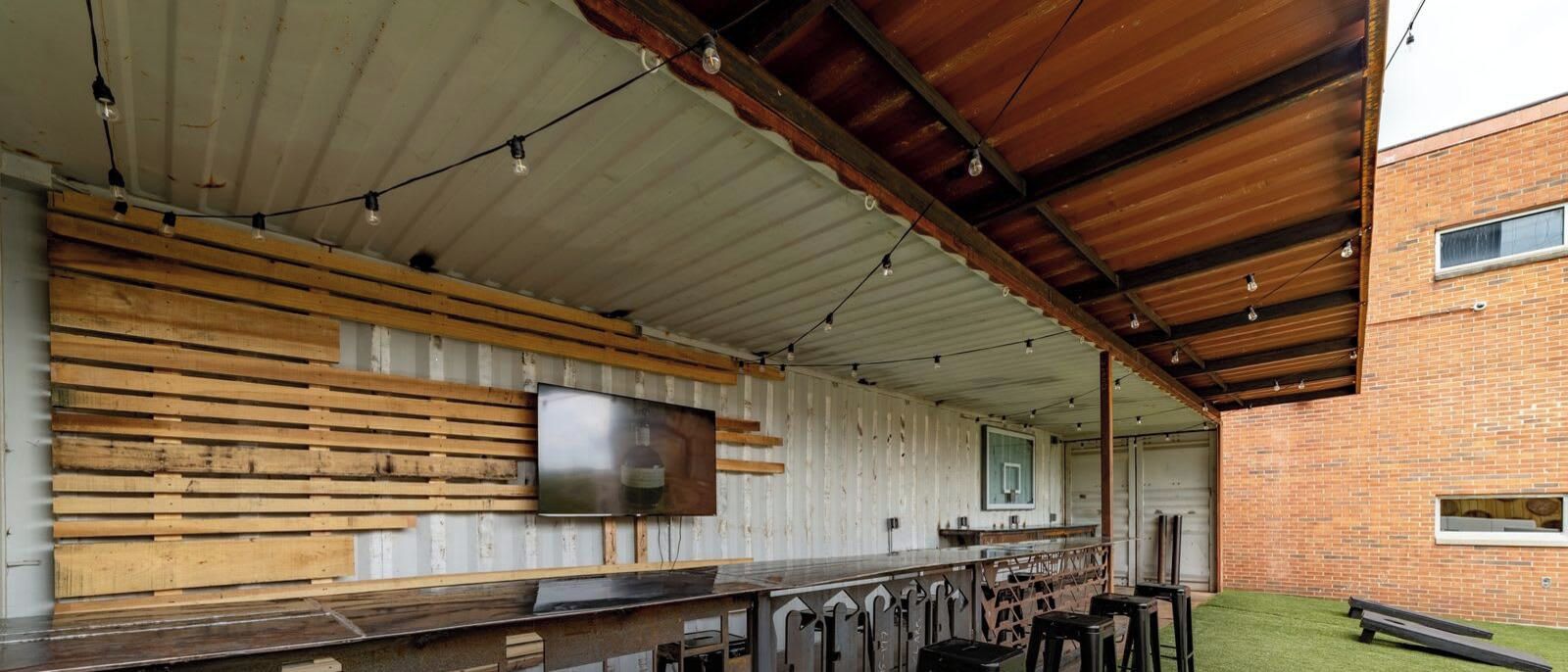 Uma área de bar ao ar livre com um balcão comprido, ripas de madeira e uma televisão fixada na parede.