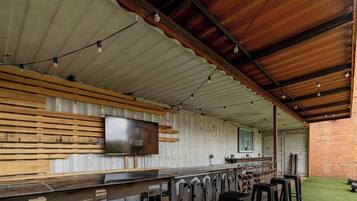Uma área de bar ao ar livre com um balcão comprido, ripas de madeira e uma televisão fixada na parede.
