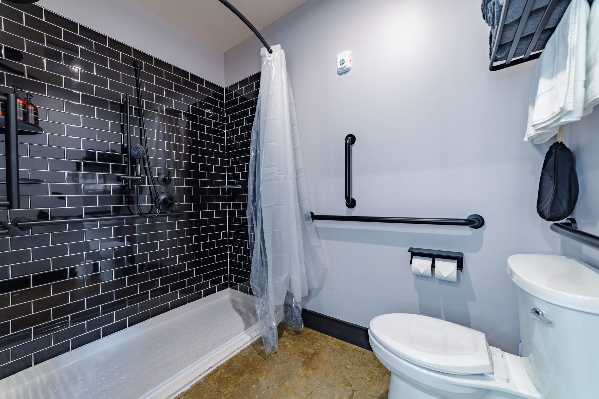 Un baño con ducha de azulejos negros, inodoro blanco, toallero y barra de apoyo.