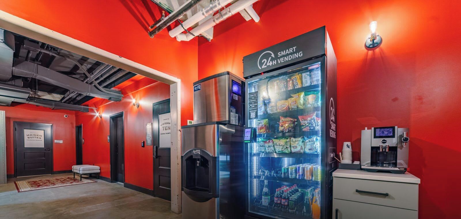 Máquina automática de venda com indicação de serviço 24 horas, um dispensador de água e uma máquina de café, numa divisão com paredes vermelhas e uma porta visível identificada como 'MAIDEN SUITES'.