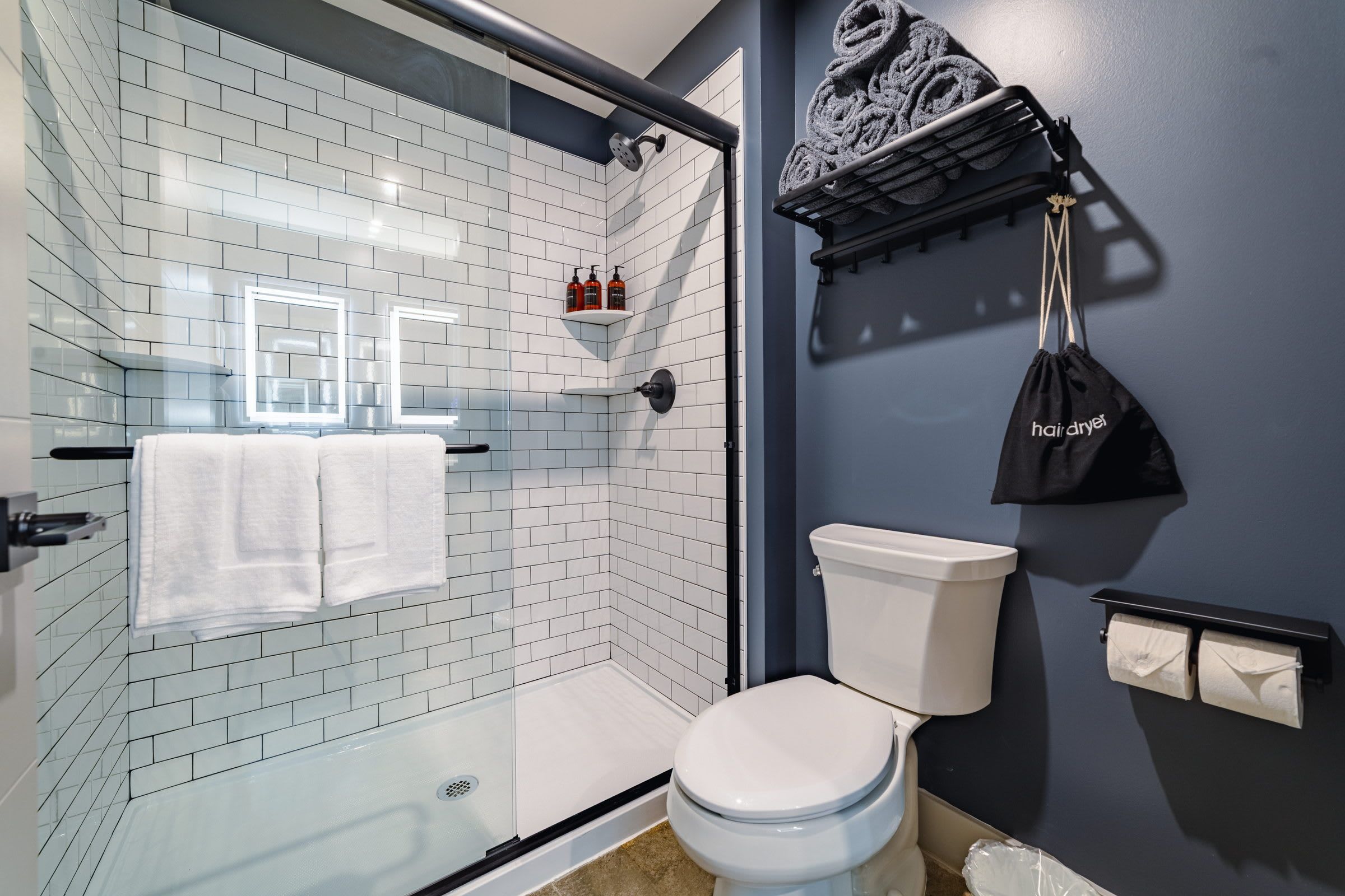 Un baño con cabina de ducha de vidrio, paredes de azulejos blancos, inodoro blanco y un perchero con secador de pelo.