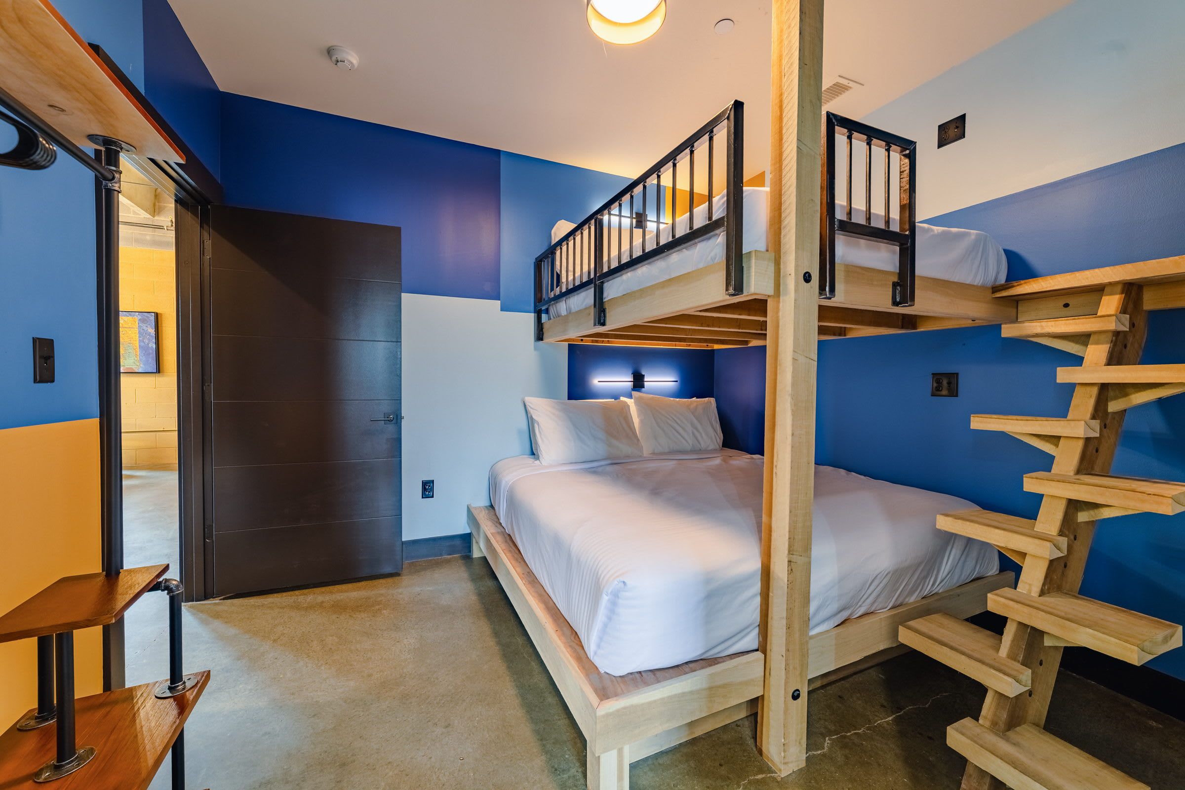 Une chambre avec un lit superposé, une échelle en bois, une porte et un luminaire fixé au mur.