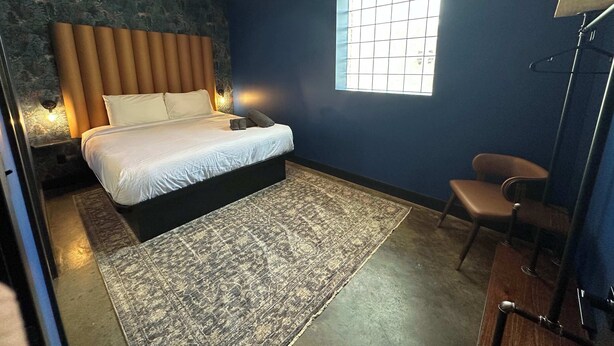 Een slaapkamer met een bed, een raam, een stoel en een nachtkastje met een lamp.