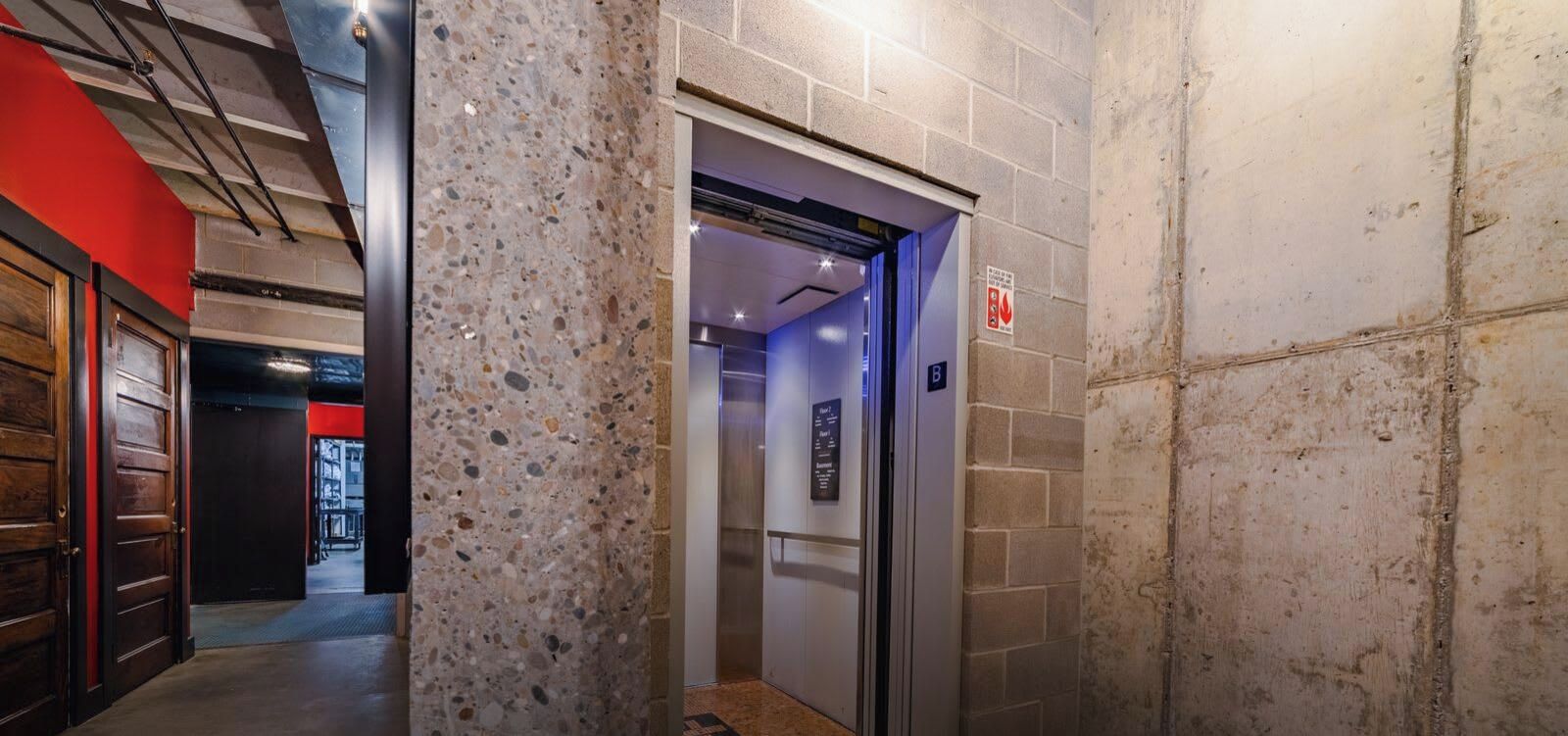Ascensore con indicazione del piano, cartello di sicurezza antincendio e corridoio con parete rossa e una porta.