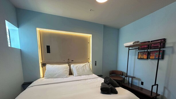 Een hotelkamer met een bed, een stoel, een nachtkastje, een wandplank met kunst en een plafondlamp.