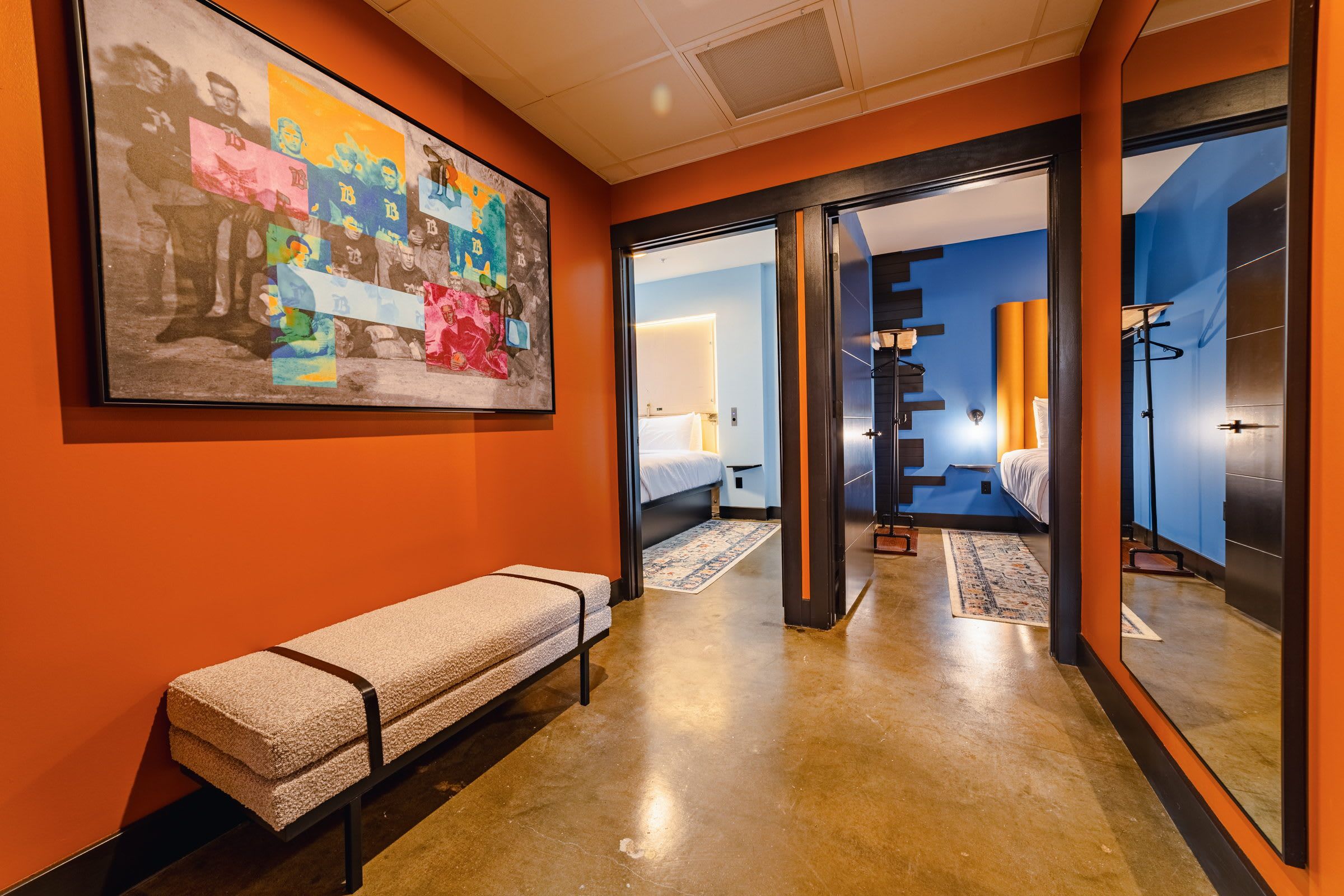 Ein Hotelgang mit einer Bank, einem großen, farbenfrohen Kunstwerk an der Wand und einem Blick in ein Schlafzimmer mit Bett und Nachttisch.