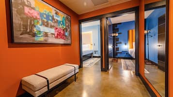 A hotel corridor with a bench, a large colorful artwork on the wall, and a glimpse of a bedroom with a bed and a nightstand.