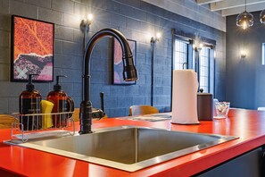 A modern kitchen with a stainless steel sink, black faucet, and a red countertop. There are dish soap and sponges on the counter, and a paper towel dispenser is visible. - Sleep48 24Beds 16BR Super Stays A12 Pad Superfecta (Louisville)