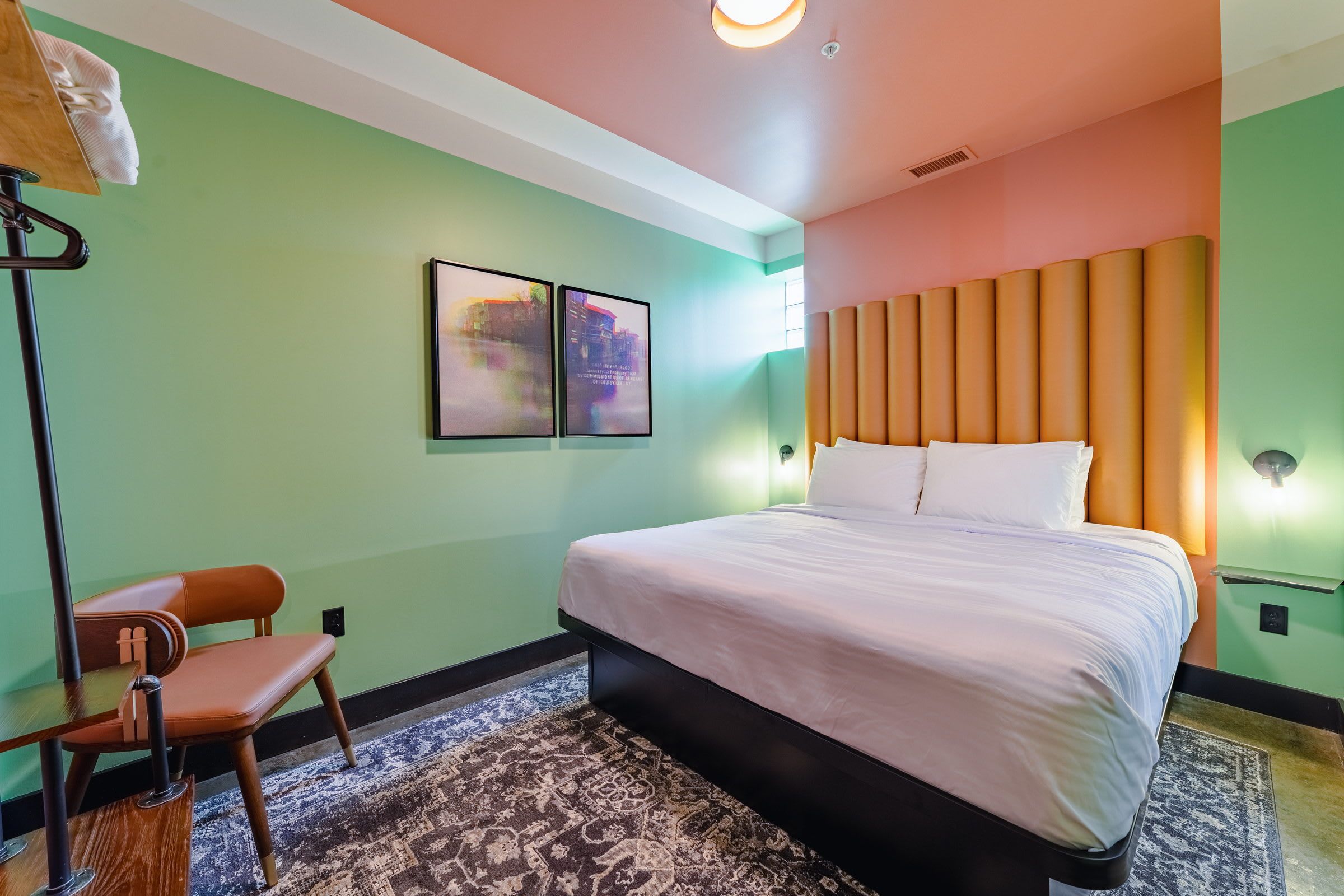 Una camera d'albergo con un letto, una sedia, un tappeto e due quadri appesi al muro.