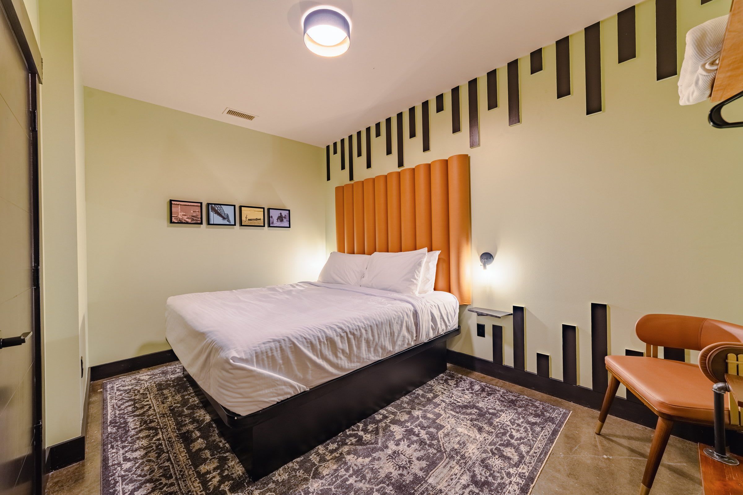 A hotel room with a bed, a chair, a rug, a wall with pictures, and a ceiling light.