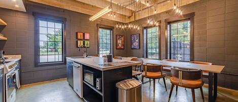 A modern kitchen with a central island, built-in oven, and a sink. There are large windows providing natural light and views of the outdoors.