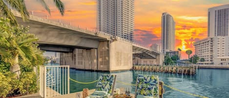 A poolside area with lounge chairs, a dining table, and a view of a city skyline at sunset.