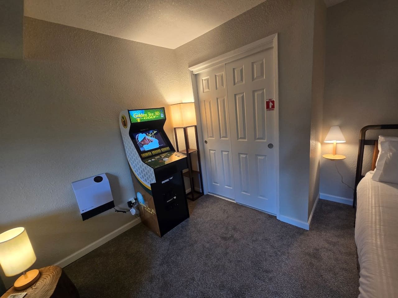 Quarto de hotel com console de jogos, porta, luminária e cama.