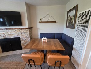 Uma sala de estar aconchegante com lareira de pedra, uma mesa de jantar de madeira com duas cadeiras, um sofá azul e uma televisão na parede.
