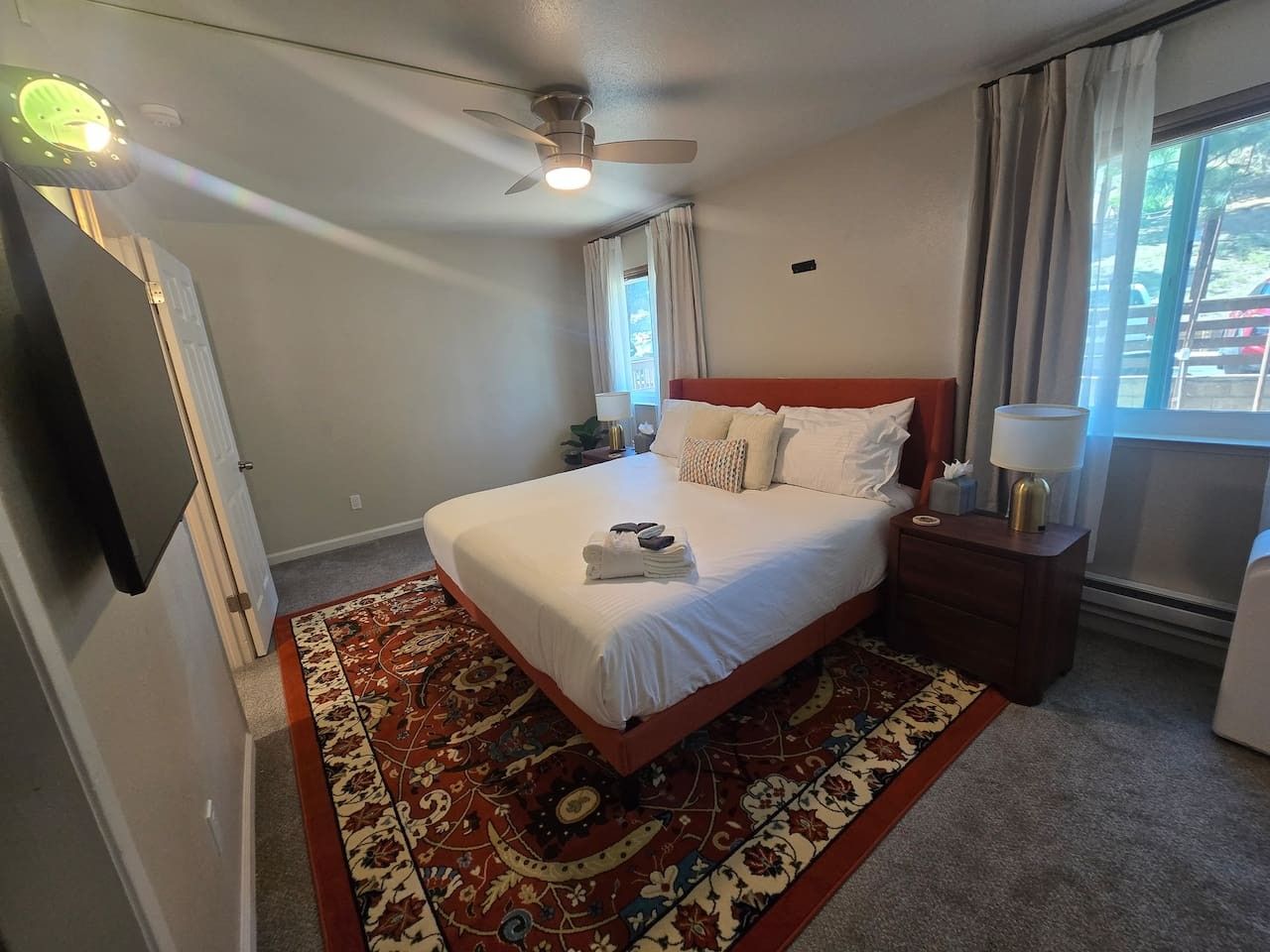 Quarto de hotel com cama, mesa de cabeceira, luminária, ventilador de teto e janela com cortinas.