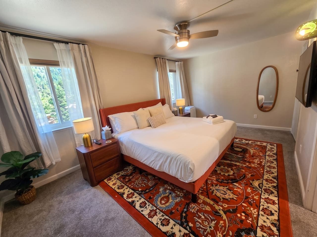 Une chambre à coucher comprenant un lit, des tables de chevet, un ventilateur de plafond, une fenêtre avec des rideaux, un miroir et un tapis à motifs.