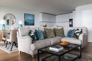 A modern living room with a beige sofa, a glass coffee table, and a dining area with a chair and table. - Liner Apartment 44 - 3 Bedroom - Fourth Floor (Falmouth)
