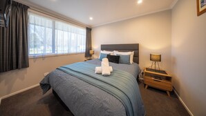 Un dormitorio con cama, mesita de noche, lámpara y ventana con cortinas.