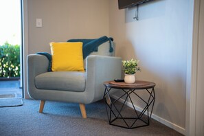 Ein modernes Wohnzimmer mit einem grauen Sofa, einem gelben Kissen, einem kleinen runden Tisch mit einer Pflanze und einem an der Wand montierten Fernseher.