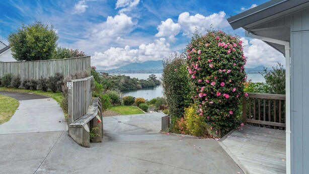 A residential property with a concrete driveway leading to a wooden deck, a fenced garden area, and a view of a lake with mountains in the distance.