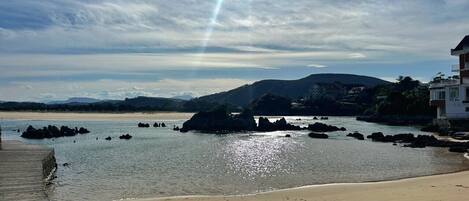 A beach with a wooden walkway, rocky outcrops in the water, and a mountain in the background.