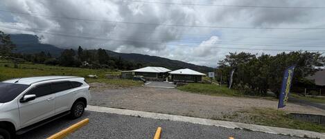 A parking area with a white SUV, a gravel surface, and a building with a green roof in the background.