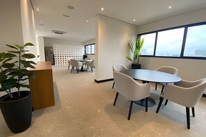 A modern office space with a round table and chairs, a potted plant, and a view through large windows. - Studio com sacada na Paulista  (São Paulo)