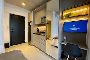 A modern, compact hotel room with a kitchenette, a bed, a desk with a chair, and a TV displaying a 'Check In' screen. - Studio com sacada na Paulista  (São Paulo)