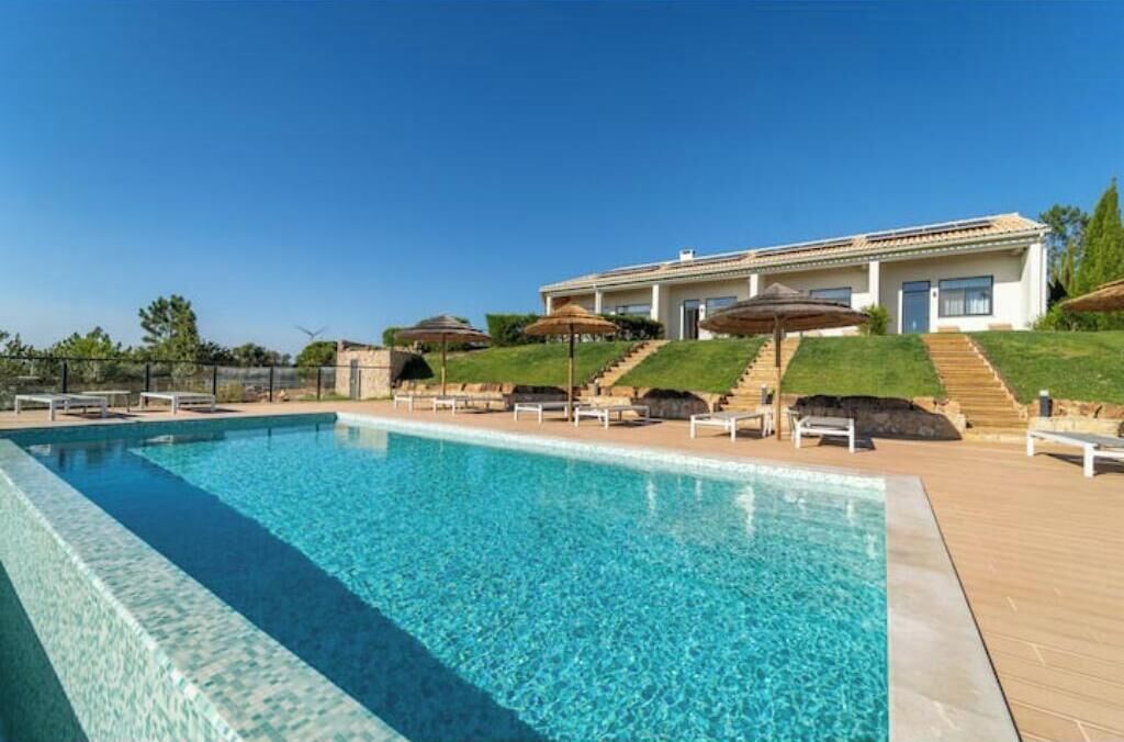 A swimming pool with a clear blue water surface, surrounded by a wooden deck, and a building with a white facade and a sloped roof in the background.
