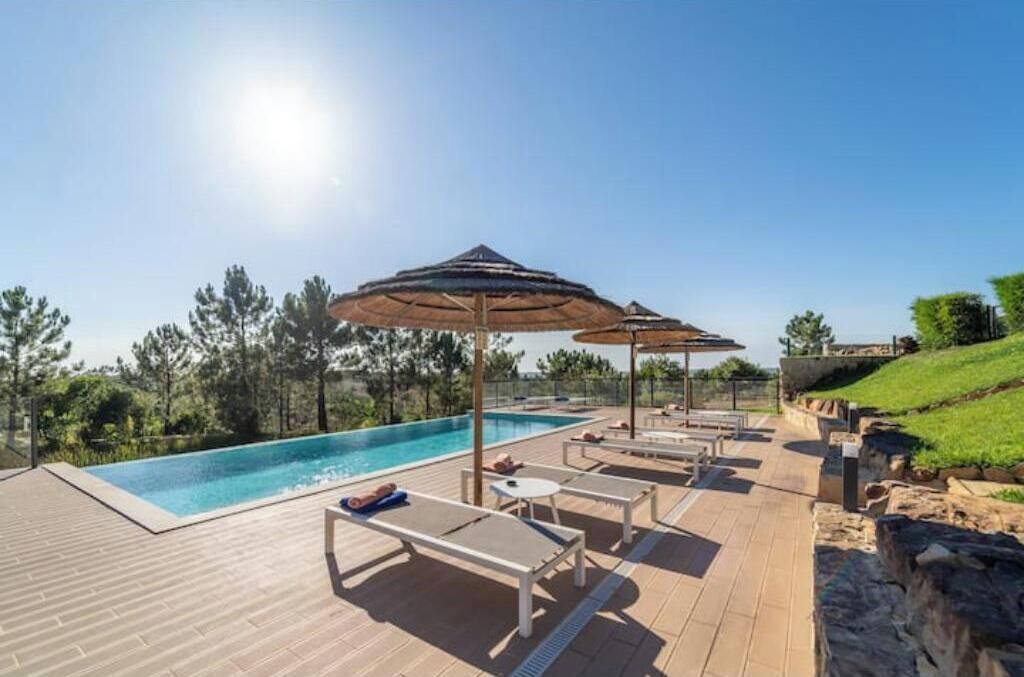Uma área de piscina com espreguiçadeiras e guarda-sóis, cercada por vegetação e um céu azul.