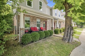 A two-story residential building with a brick facade, a covered porch with red chairs, and a well-maintained lawn.