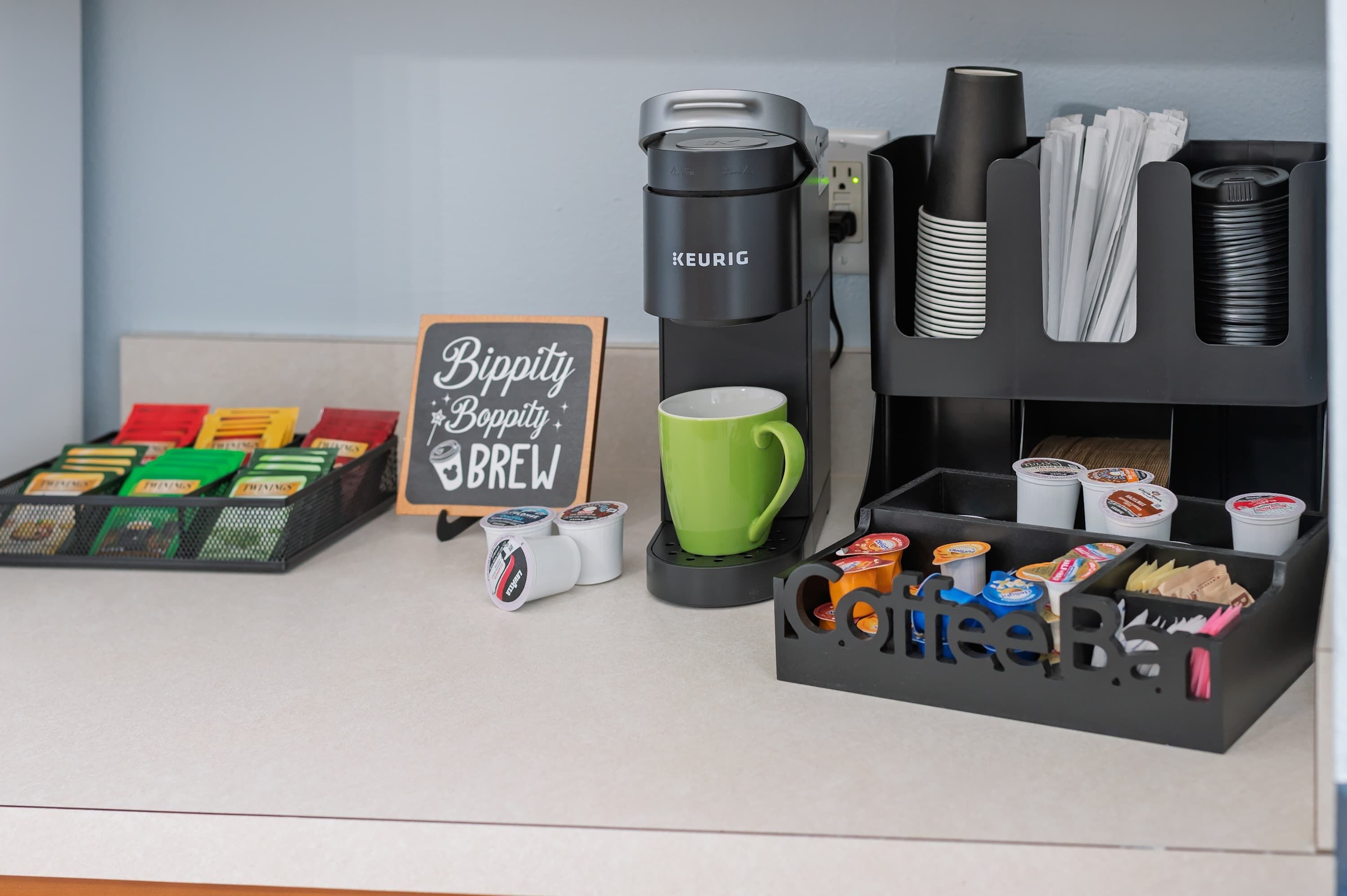 A coffee station with a Keurig coffee maker, a green mug, and a sign that reads 'Bippity Boppity Brew.'