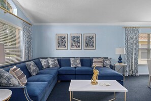 Een woonkamer met een blauwe bank, een witte salontafel en decoratieve kussens. Drie ingelijste plantendrukken hangen aan de muur.