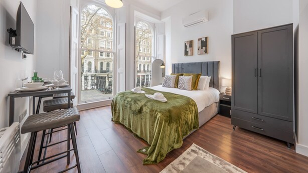 Ein Schlafzimmer mit einem großen Bett, einem Kleiderschrank, einem Schreibtisch mit Stuhl und Blick auf Gebäude durch ein Fenster.