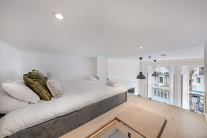 Ein Schlafzimmer mit Bett, Kissen, Nachttisch, einem Fenster mit Blick auf Gebäude und einem Balkon.