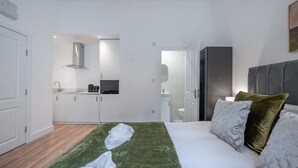 Een hotelkamer met een bed, een groene deken, een grijs hoofdeinde en een kitchenette op de achtergrond.