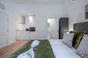 Ein Hotelzimmer mit einem Bett, einer grünen Decke, einem grauen Kopfteil und einer Küchenzeile im Hintergrund.