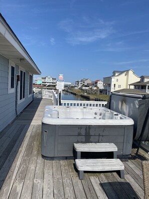 A wooden deck with a hot tub and a small bench, overlooking a water body and residential buildings. - Canalfront dock, boat ramp and hot tub (North Topsail Beach)