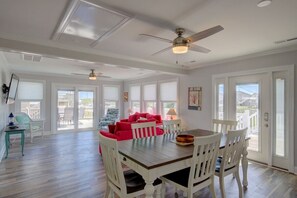 A spacious dining area with a wooden table and white chairs, a ceiling fan, and large windows with blinds. - Canalfront dock, boat ramp and hot tub (North Topsail Beach)