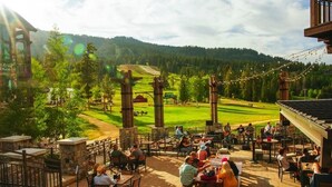 An outdoor dining area with tables and chairs, overlooking a golf course and forested hills. - Tamarack Resort Village Ski In Ski Out Condo - Year Round Resort (Donnelly)