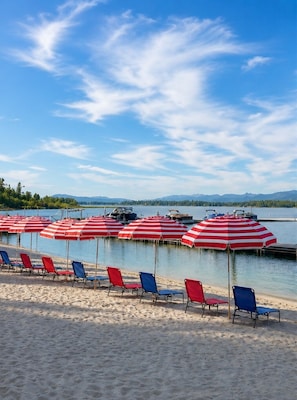 Ein Strand mit Reihen aus gestreiften Sonnenschirmen und Liegestühlen, ein ruhiger See und ferne Berge.