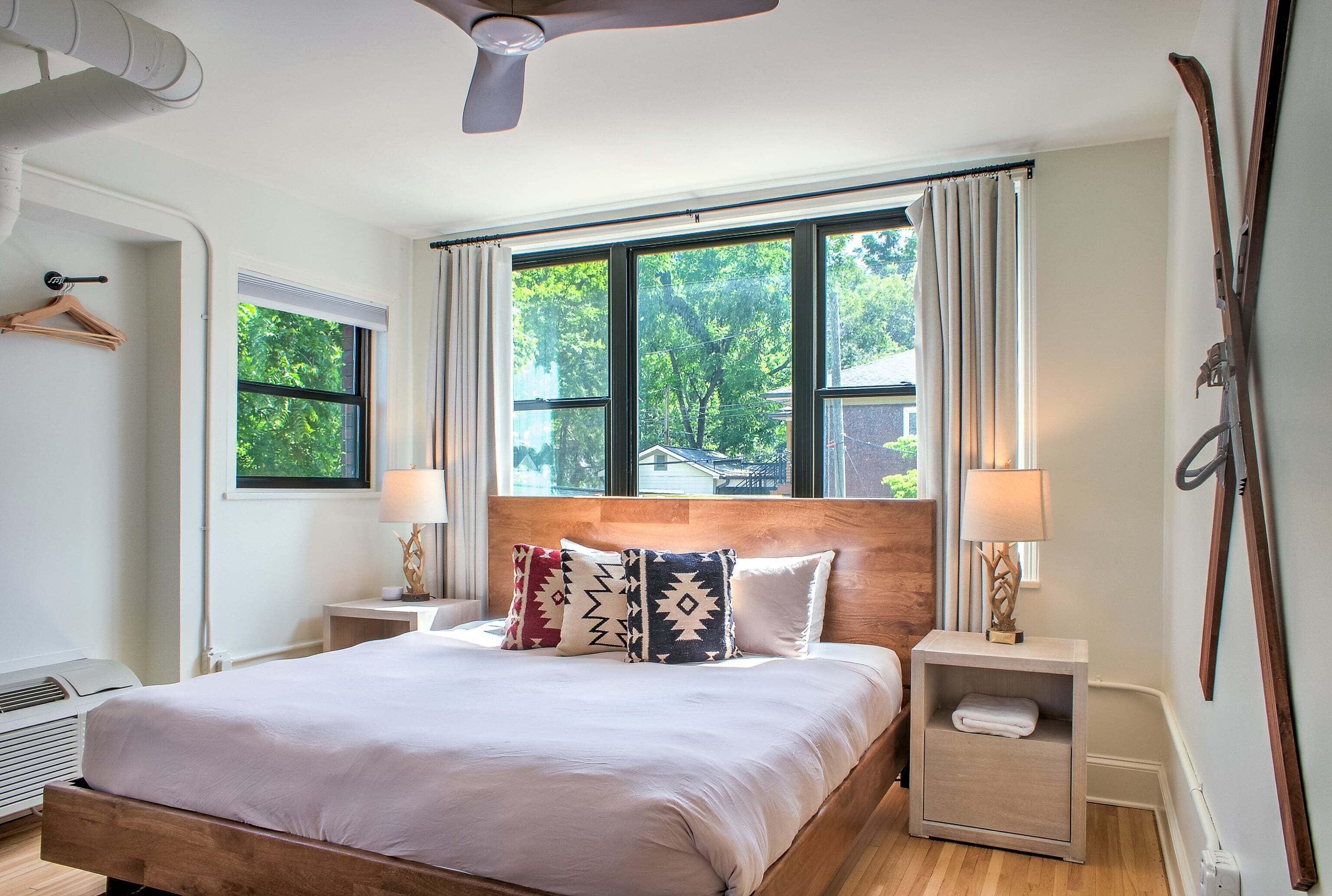Ein Hotelzimmer mit einem Bett, zwei Nachttischen, einem Fenster mit Vorhängen, einem Deckenventilator und einem Skiständer.