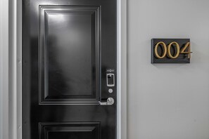 Eine schwarze Tür mit einem Zahlenschloss und einem kleinen Fenster. Dazu eine schwarze Schild mit der Nummer '004' in Gold.