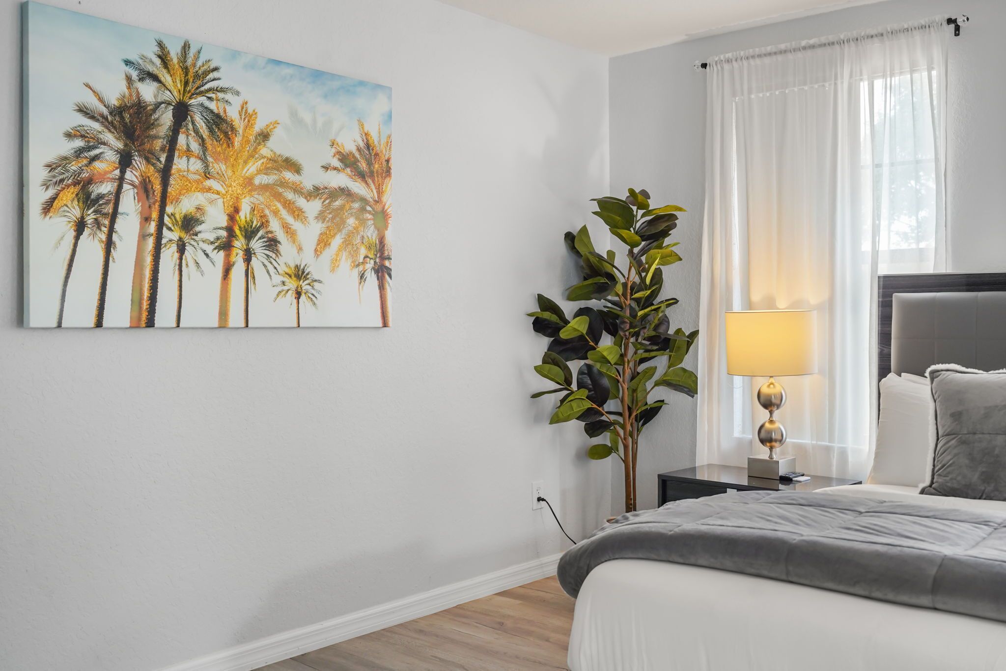 Ein Schlafzimmer mit einem Bett, einem Nachttisch, einer Lampe, einer an der Wand befestigten Pflanze und einem Palmen-Gemälde.